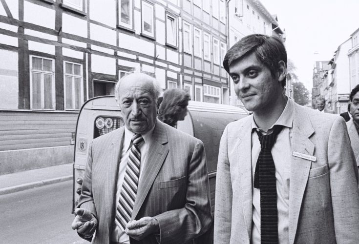 Schwarzweißfoto von zwei Männern, die auf einer Straße entlanggehen, mit historischen Gebäuden im Hintergrund.