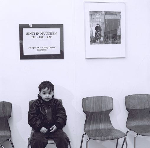 Ein Junge sitzt alleine auf einem Stuhl vor einer Wand mit einer Ausstellungstafel und einem Schwarzweißfoto einer Sinti-Frau.