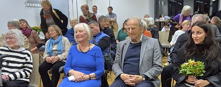 Ein Publikum sitzt aufmerksam bei der Vernissage von Milly Orthens Ausstellung "Sinti und Roma, gestern und heute" im Neuhauser Trafo. Die Anwesenden sind älteren und jüngeren Alters und zeigen Interesse an den präsentierten Fotografien.