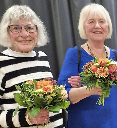 Zwei ältere Frauen lächeln in die Kamera, während sie bunte Blumensträuße halten. Die Frau links trägt ein gestreiftes Oberteil und Brille, die Frau rechts trägt ein blaues Kleid.