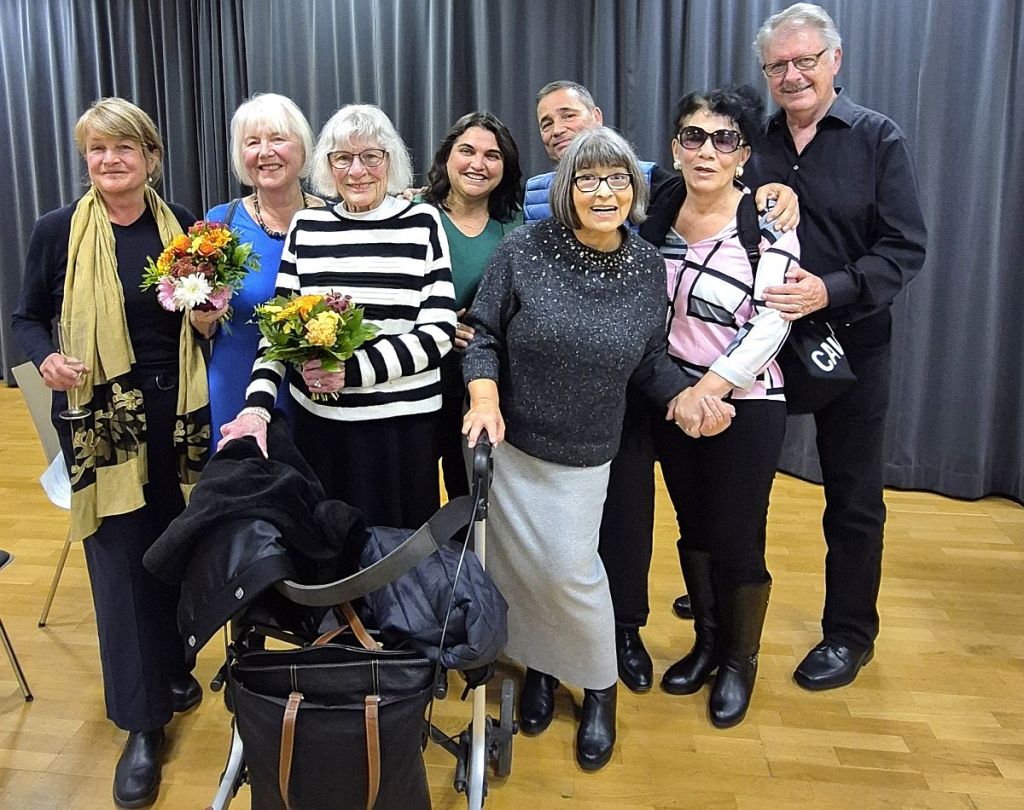 Eine Gruppe von Menschen steht zusammen und lächelt in die Kamera, einige halten Blumen in den Händen. Im Hintergrund ist ein dunkler Vorhang zu sehen.