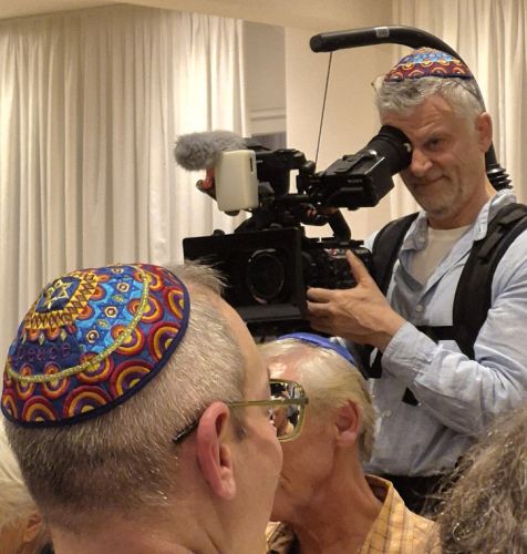 Ein Mann mit einer Kamera filmt eine Gruppe von Menschen, die bunte Kippot tragen. Die Kamera wird auf den Mann mit dem grauen Haar gerichtet, der einen interessierten Ausdruck zeigt.