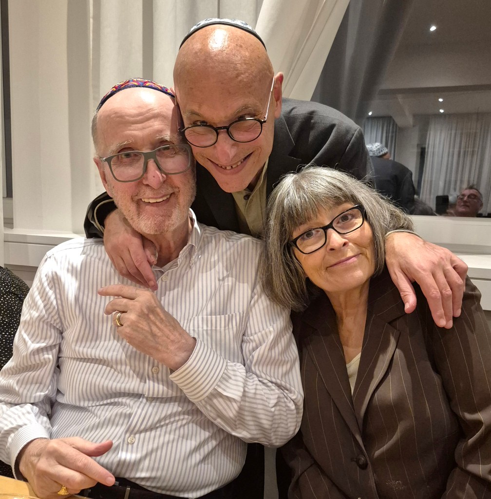 Drei Personen posieren für ein Foto in einem hellen Raum. Der Mann in der Mitte trägt eine Brille und lächelt, während er seine Arme um die anderen beiden legt. Eine Frau mit grauen Haaren und einer Brille sitzt rechts, während ein weiterer Mann links steht und lächelt.
