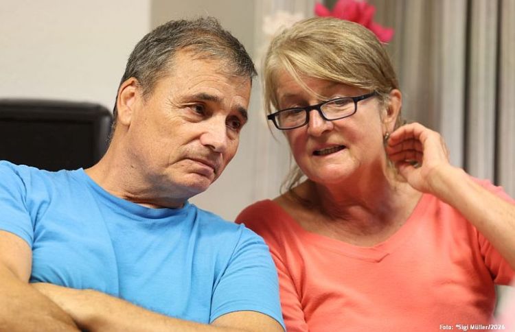 Ein Mann und eine Frau sitzen eng beieinander und führen ein Gespräch. Der Mann trägt ein blaues T-Shirt und schaut nachdenklich, während die Frau in einem roten T-Shirt mit Brille spricht und ihn aufmerksam betrachtet.