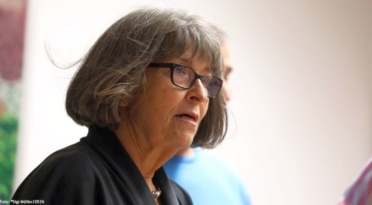 Eine Frau mit grauen Haaren und Brille spricht nachdenklich. Im Hintergrund sind verschwommene Personen sichtbar.
