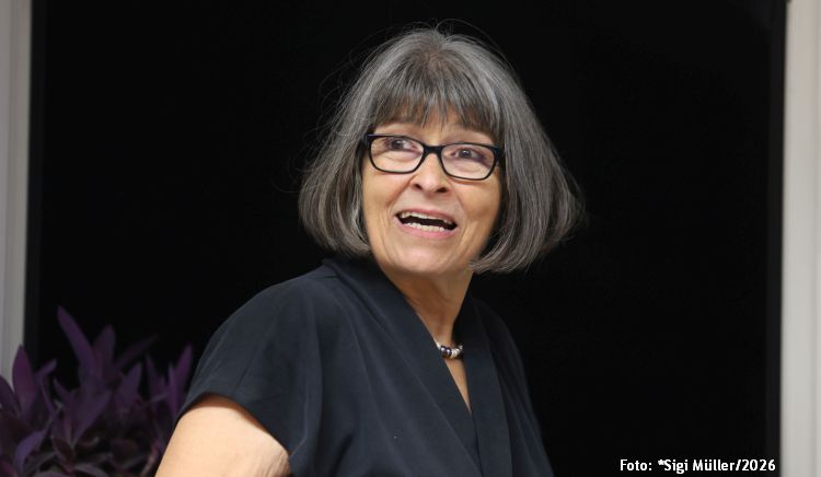 Lächelnde Frau mit grauen Haaren und Brille, trägt ein schwarzes Oberteil, vor einem dunklen Hintergrund.
