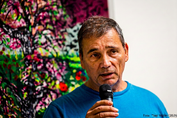 Ein Mann mit einem Mikrofon spricht, im Hintergrund ist ein buntes, gemaltes Kunstwerk zu sehen.