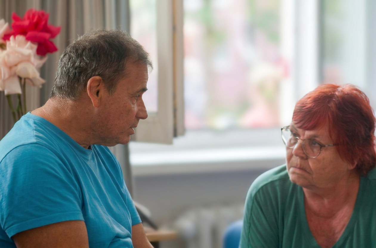 Ein Mann und eine Frau sitzen sich gegenüber und führen ein ernstes Gespräch. Im Hintergrund sind Blumen und Fenster zu sehen.