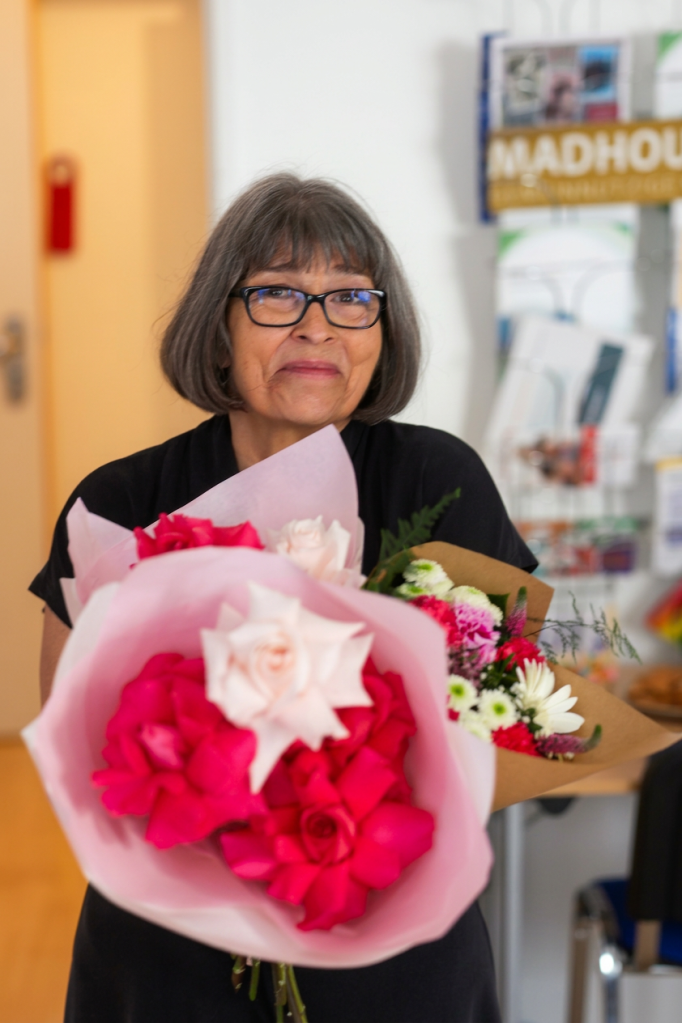 Eine lächelnde Frau mit Brille hält zwei Blumensträuße in Händen, die rosafarbene und rote Rosen sowie andere bunte Blumen enthalten. Im Hintergrund ist ein heller Raum zu sehen.