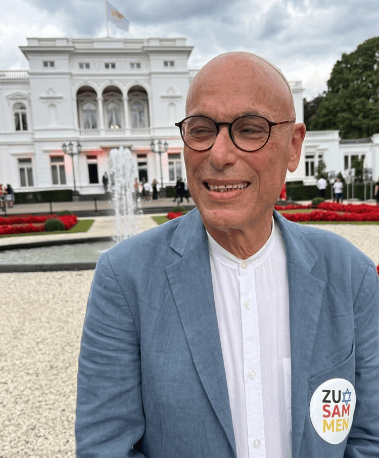 Ein älterer Mann mit Brille lächelt vor einem historischen Gebäude mit weißen Wänden und Säulen. Blumenbeete und ein Springbrunnen sind im Hintergrund sichtbar. Der Mann trägt einen hellblauen Blazer und ein weißes Hemd, sowie einen Aufkleber mit der Aufschrift 'ZUSAMMEN'.