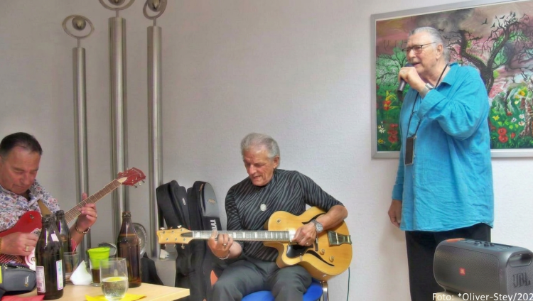Drei Männer spielen Musik in einem Raum; einer singt, während die anderen Gitarre spielen. Im Hintergrund sind Flaschen und ein buntes Gemälde zu sehen.