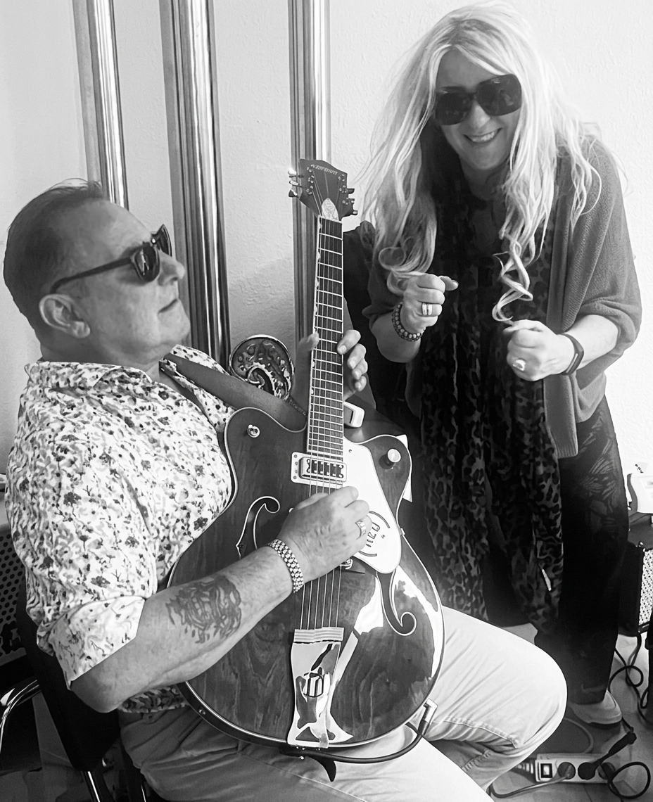 Ein Mann sitzt auf einem Stuhl und spielt eine Gitarre, während eine Frau mit langen Haaren und Sonnenbrille daneben steht und fröhlich tanzt.