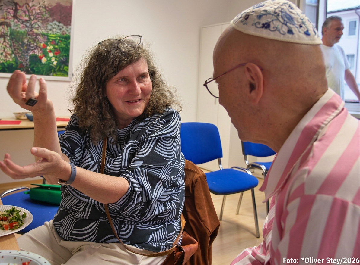 Eine Frau mit lockigem Haar und einer gestreiften Bluse spricht mit einem Mann, der eine Kippa trägt. Sie gestikuliert mit der Hand, während sie an einem Tisch mit Essen sitzt. Im Hintergrund sind blaue Stühle und ein Bild an der Wand zu sehen.