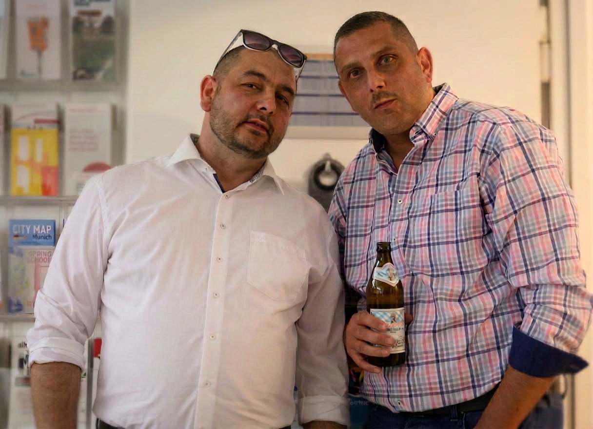 Zwei Männer posieren in einem Innenraum, einer mit Sonnenbrille und der andere hält eine Flasche Bier.