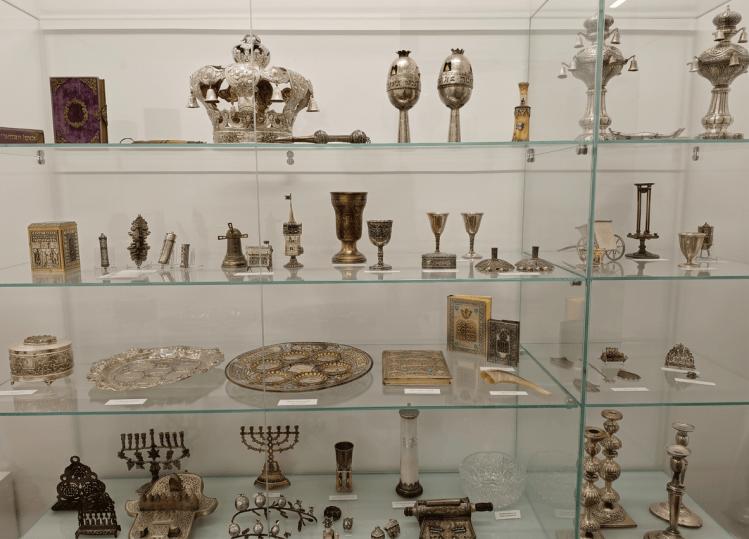 Ein Glasvitrinen mit einer Sammlung von silbernen religiösen Gegenständen, darunter eine Krone, Kelche, Menorahs und weitere verzierte Objekte.