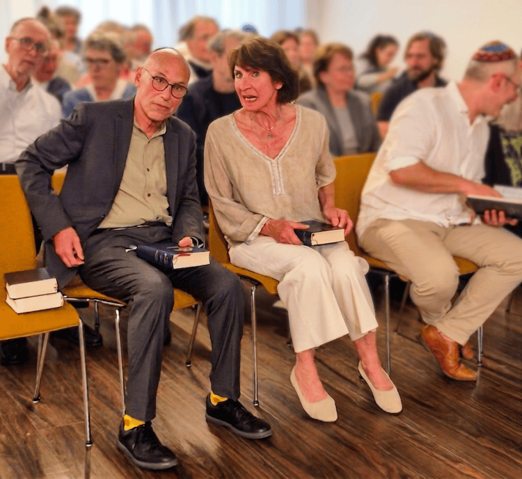 Zwei Personen sitzen auf Stühlen in einem Raum voller Zuschauer. Sie halten Bücher in den Händen und schauen aufmerksam. Der Mann trägt eine graue Jacke und eine Brille, die Frau hat ein helles Oberteil an. Im Hintergrund sind weitere Teilnehmer sichtbar.