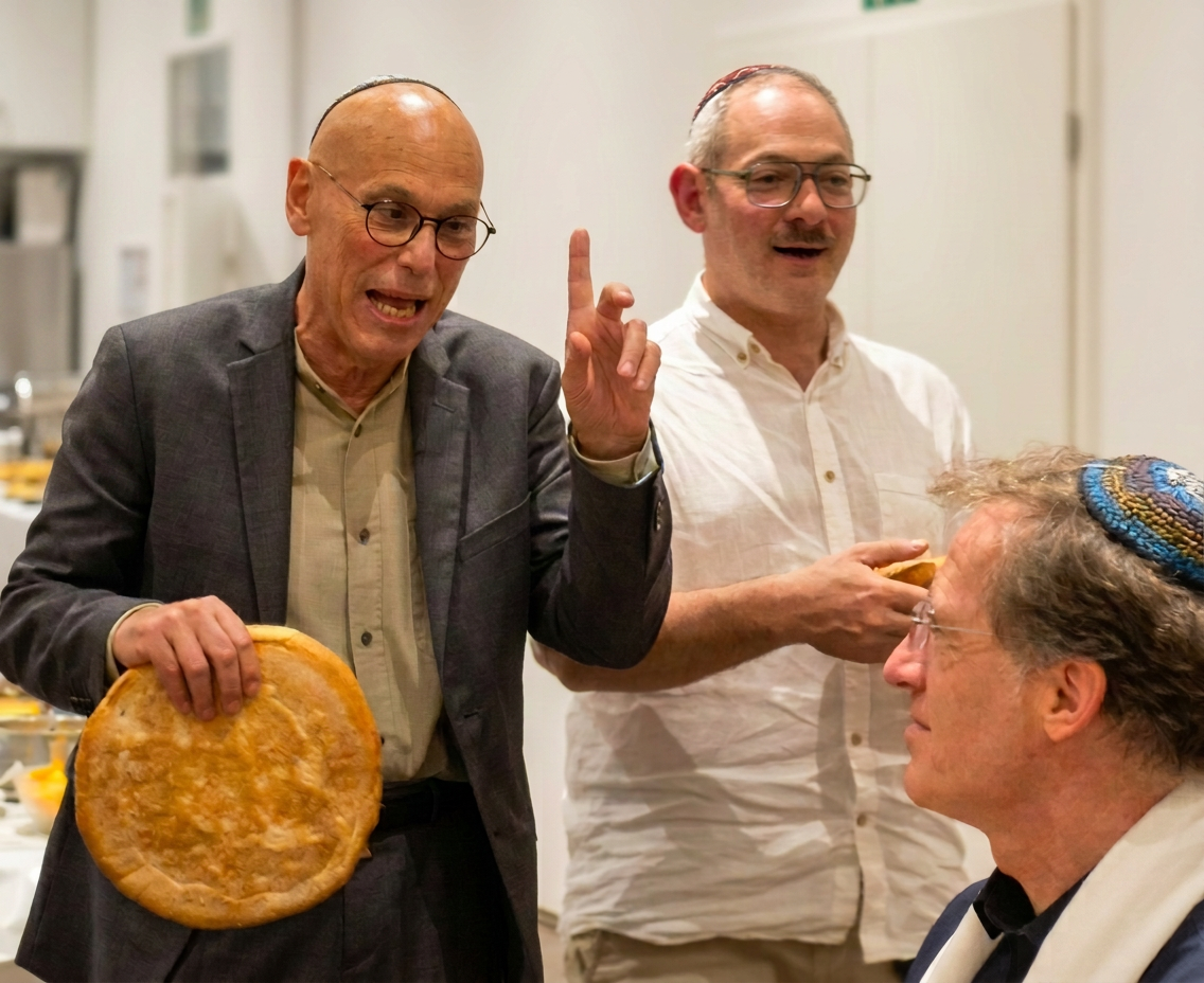 Drei Männer in formeller Kleidung sprechen und lachen bei einer Zusammenkunft, einer hält einen großen, runden Brotlaib in der Hand.