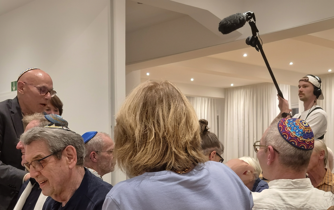 Eine Gruppe von Menschen in einem Raum, einige tragen Kippahs, während ein Mann im Hintergrund ein Mikrofon hält.