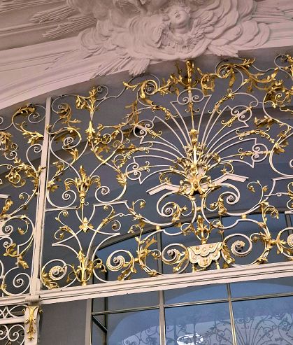 Detailaufnahme eines eleganten, goldenen Metallgitter mit floralen Motiven und geschwungenen Linien, vor einem blauen Hintergrund.