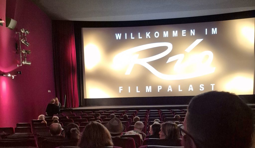 Einblick in ein Kino mit dem Schild 'Willkommen im Río Filmpalast' auf der Leinwand, rote Sitze und Zuschauer im Vordergrund.
