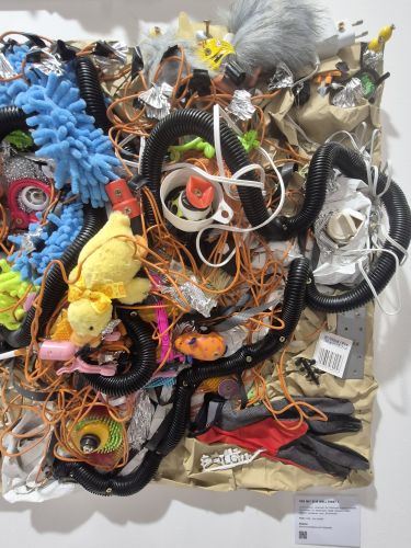 Ein buntes Kunstwerk aus verschiedenen Objekten und Materialien, darunter Spielsachen, Verpackungsmaterial und Kabel, die kreativ angeordnet sind.