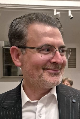 Ein lächelnder Mann mit Brille und grauen Haaren, der einen Anzug trägt.
