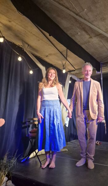 Zwei Personen stehen auf einer Bühne, halten Händchen und lächeln. Die Frau trägt ein weißes Oberteil und einen blauen Rock, der Mann ist in einem beigen Anzug gekleidet. Die Kulisse ist mit Lichtern und einem dunklen Vorhang dekoriert.