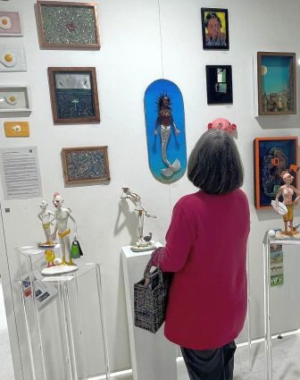 Eine Person mit grauen Haaren, die auf einer Ausstellung steht und Kunstwerke an einer weißen Wand betrachtet. Sie trägt einen roten Pullover und hält eine Tasche in der Hand. Um sie herum sind verschiedene Kunstwerke und Skulpturen zu sehen.