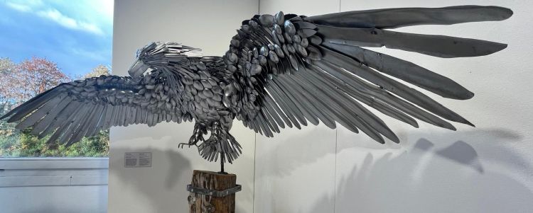Eine kunstvolle Skulptur eines Adlers aus Metallbesteck mit weit ausgebreiteten Flügeln, auf einem Holzsockel montiert.