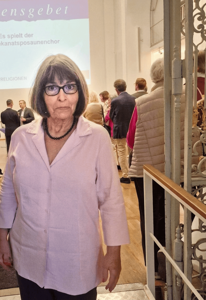 Eine ältere Frau mit Brille und grauen Haaren steht im Vordergrund einer Veranstaltung, während hinter ihr eine Gruppe von Personen in einem hellen Raum auf einem Stuhl wartet. Im Hintergrund sind Präsentationstexte zu sehen.