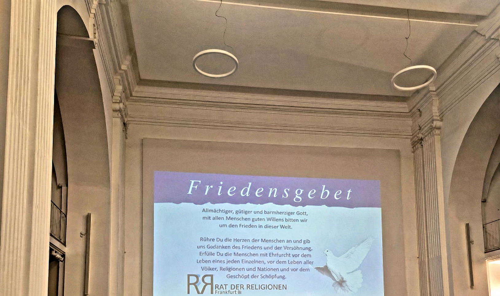 20260929_193819 Präsentation des Friedensgebets in einem großen Saal, mit einem Text auf der Leinwand und einer weißen Taube als Symbol des Friedens.