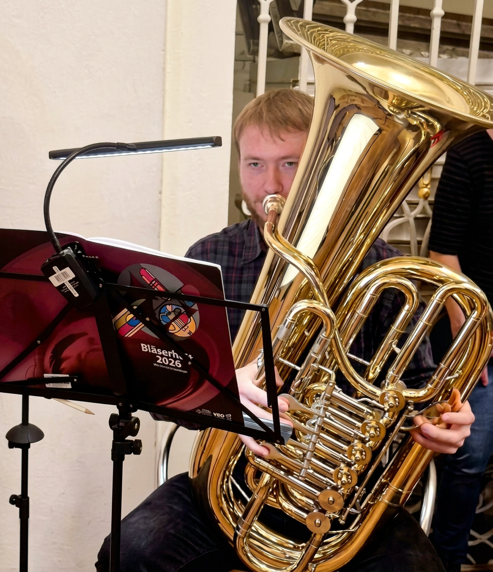 20260929_195646 Ein Mann spielt eine große Tuba während einer Probe, mit einem Notenständer vor ihm, auf dem ein Katalog für eine Veranstaltung abgebildet ist.