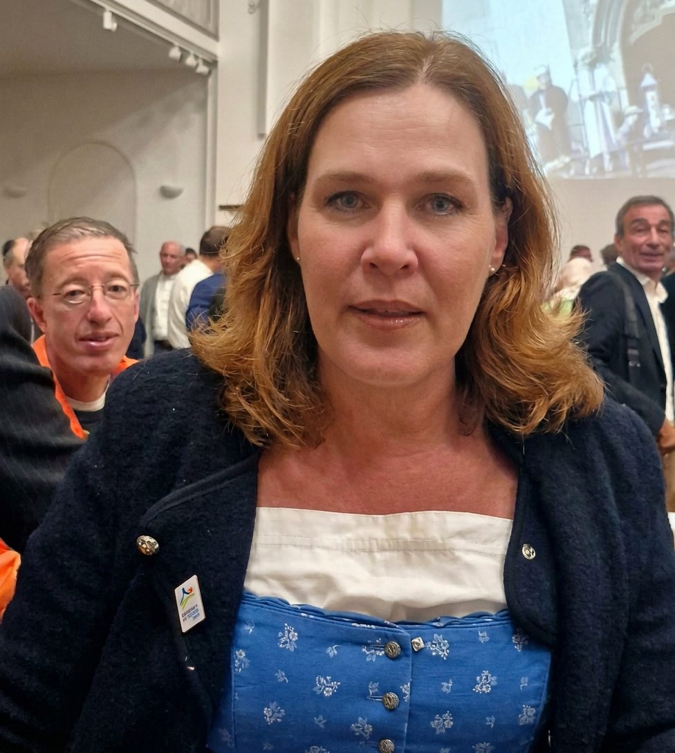 2026_09_29_Verena-Dietl_Grusswort_Friedensgebet_10-Jahre-Rat-der-Religionen_ehemalige-Karmeliterkirche-Muenchen_GdS-Blog Eine Frau mit lockigem, halblangen Haaren trägt ein traditionelles bayerisches Dirndl in Blau mit weißen Blumenmustern. Im Hintergrund sind Menschen in einer festlichen Umgebung zu sehen.