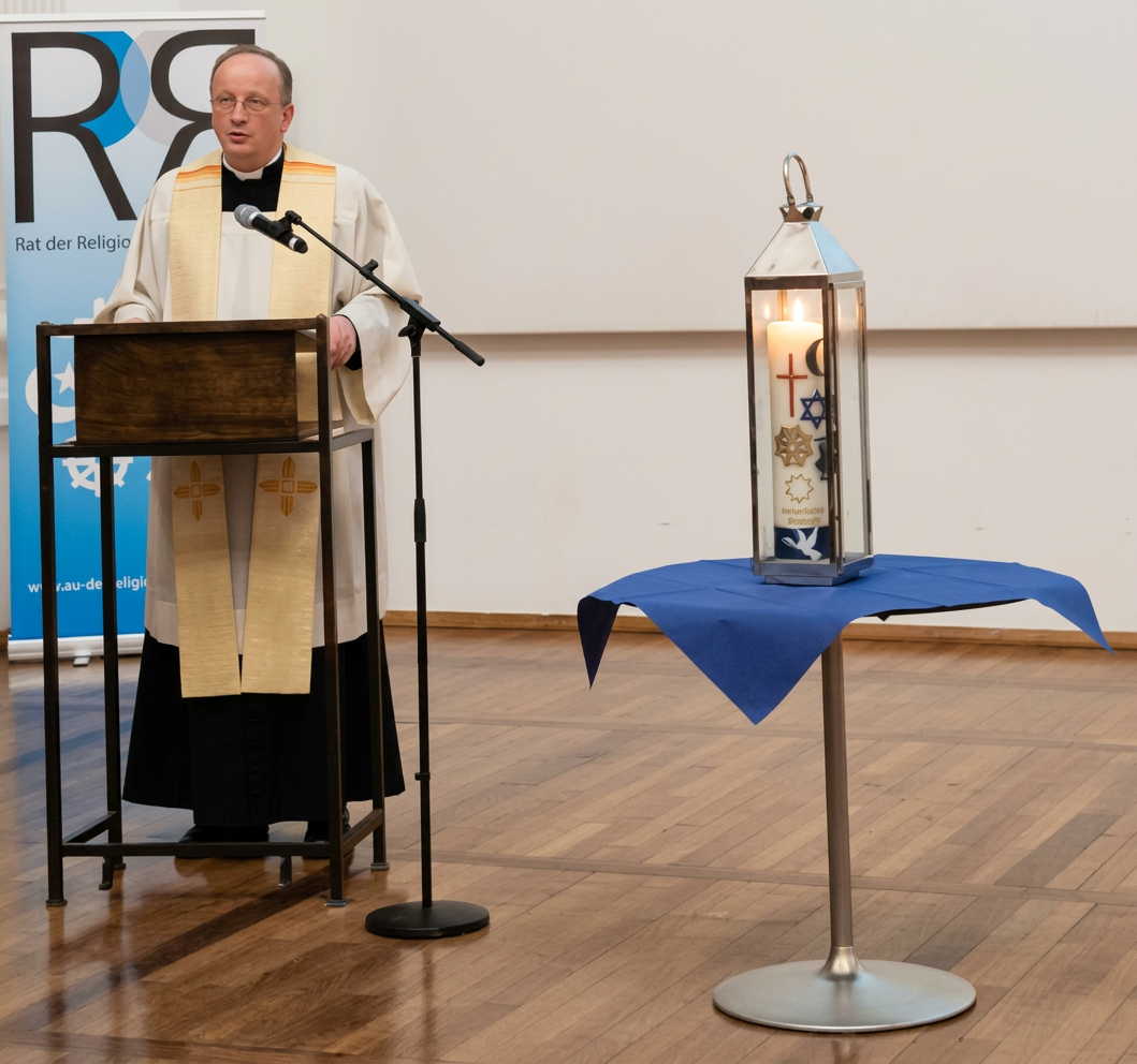 20260929_190247 Ein Redner steht an einem Mikrophon hinter einem Holzpult, während neben ihm eine Laterne mit einer brennenden Kerze auf einem Tisch steht. Die Laterne zeigt religiöse Symbole. Im Hintergrund ist ein Banner des 'Rates der Religionen' zu sehen.
