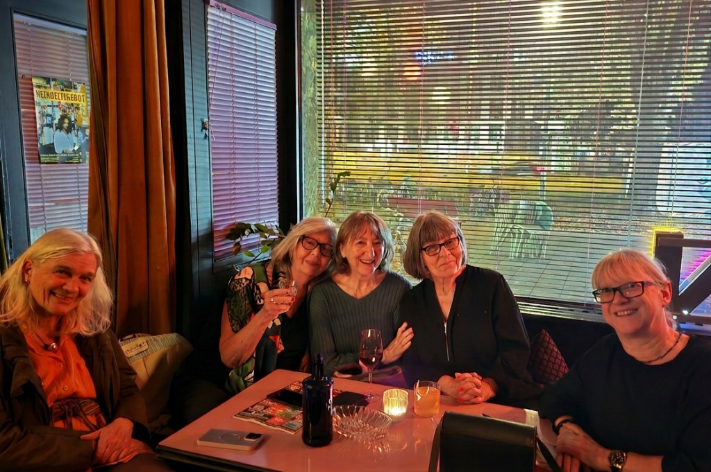 Fünf Frauen lächeln und sitzen gemeinsam in einem gemütlichen Raum mit Fenstern und sanfter Beleuchtung. Eine Frau hält ein Glas in der Hand.