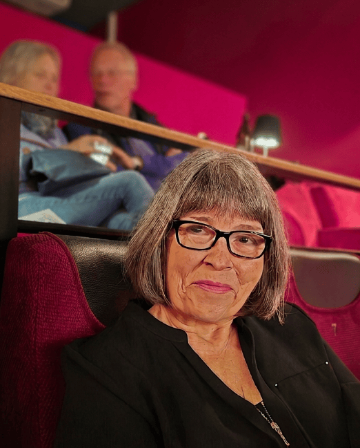 Eine ältere Frau mit grauen Haaren und Brille sitzt in einem Kino mit einer roten Polsterung im Hintergrund.