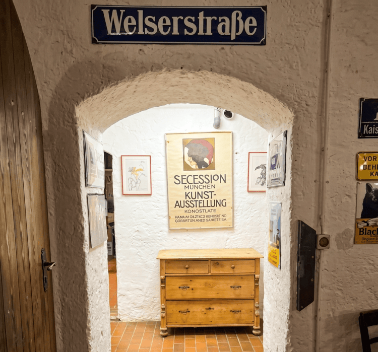 Eingang zu einem Raum mit der Aufschrift 'Welserstraße', der eine Holzkommode und mehrere gerahmte Kunstplakate zeigt.