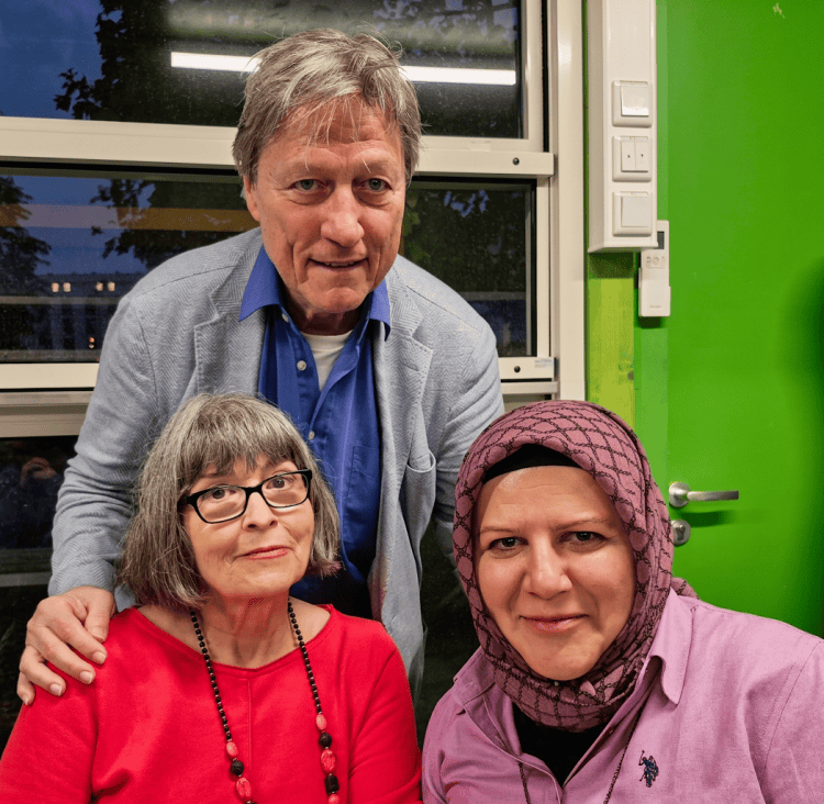 Drei Menschen posieren lächelnd vor einem Fenster. Die linke Person trägt eine graue Brille und eine rote Bluse, während die mittlere Person einen hellblauen Pulli unter einem grauen Blazer trägt. Die rechte Person hat ein Kopftuch und ein rosa Hemd an. Der Hintergrund ist grün gestrichen.