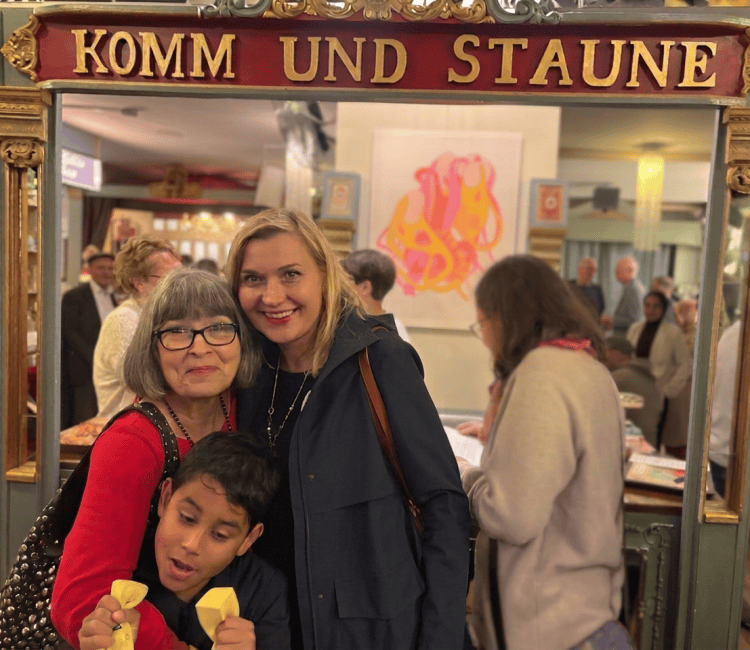 Drei Personen posieren fröhlich vor einem Schild mit der Aufschrift 'KOMM UND STAUNE', während im Hintergrund andere Gäste zu sehen sind.