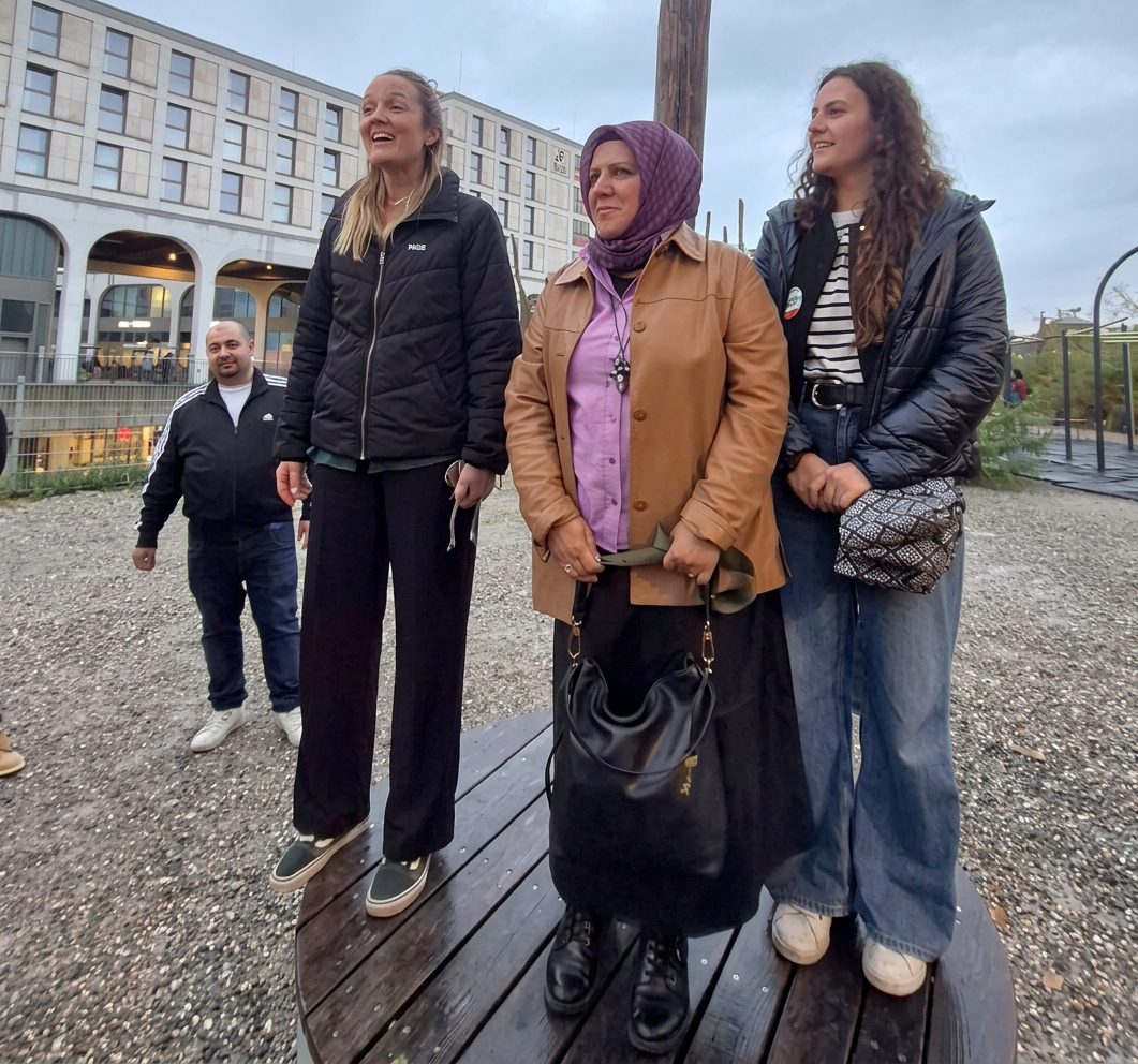 Eine Gruppe von vier Personen steht lächelnd auf einer Holzkonstruktion im Freien. Hintergrund mit moderner Architektur. Die Frauen tragen verschiedene Kleidungsstile, darunter eine Jacke und ein Kopftuch. Der Mann im Hintergrund schaut in die Kamera.
