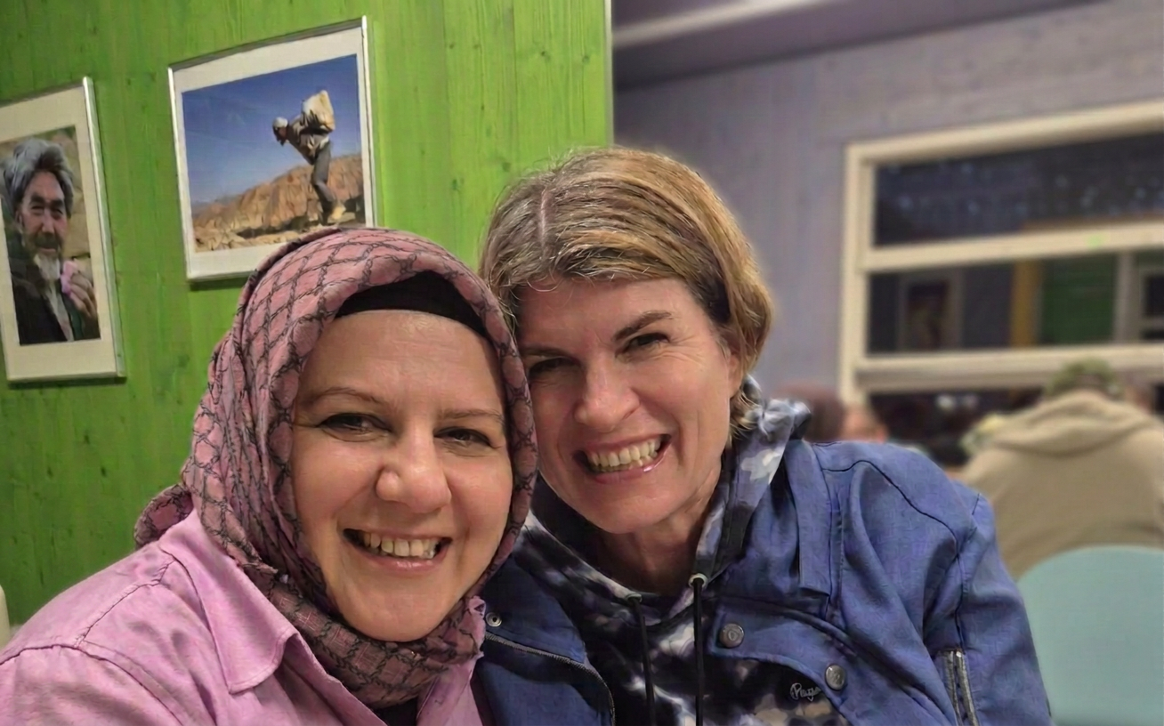 Zwei Frauen lächeln in einem Café mit grünen Wänden, eine trägt ein Kopftuch und die andere ein blaues Oberteil.