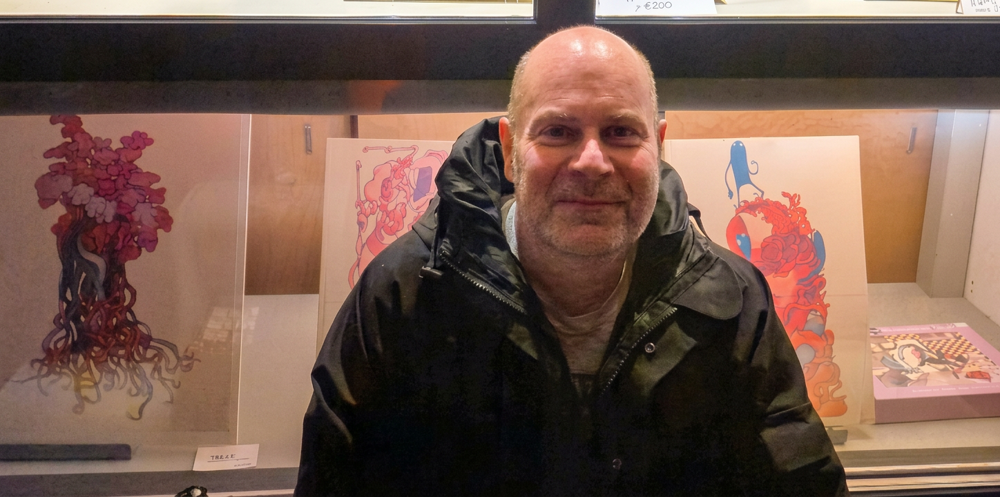 Ein Mann mit Glatze und Bart, der in einer dunklen Jacke lächelt, posiert vor einer Galerie mit bunten Kunstwerken im Hintergrund.