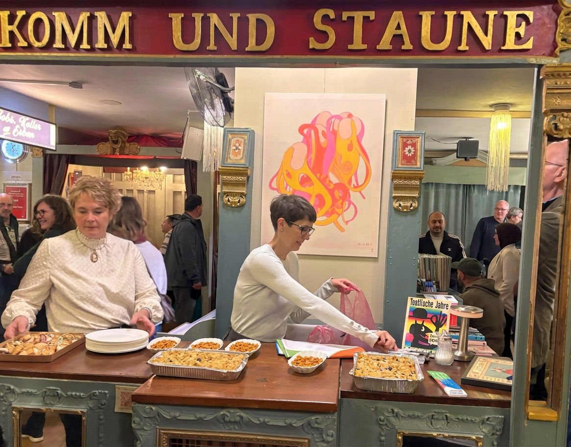 Zwei Damen bei der Zubereitung von Speisen in einem festlichen Umfeld mit Gästen im Hintergrund und einem bunten Kunstwerk an der Wand.
