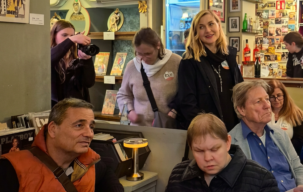 Eine Gruppe von Menschen in einem gemütlichen Raum, einige unterhalten sich, während andere miteinander interagieren. Im Vordergrund sitzt ein Mann in einem orangefarbenen Hoodie, während eine Frau im Hintergrund fotografiert. An den Wänden hängen nostalgische Bilder und Postkarten.
