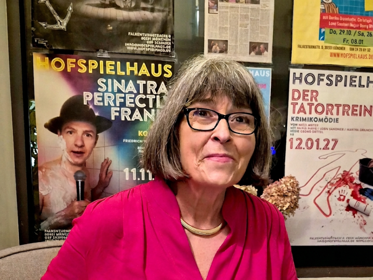 Eine Frau mit grauen Haaren und Brille, die in einem roten Oberteil lächelt, vor einer Wand mit Veranstaltungsplakaten.
