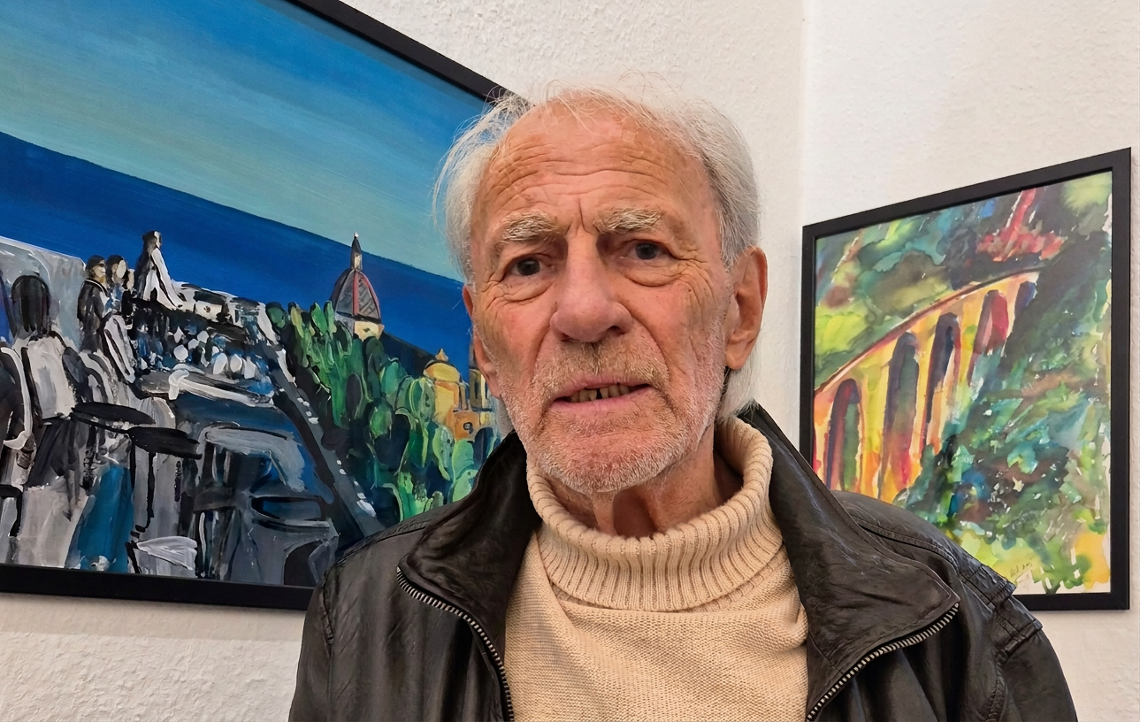 Ein älterer Mann mit grau-weißen Haaren steht in einem Raum mit zwei bunten Gemälden im Hintergrund. Er trägt eine schwarze Lederjacke und einen hellen Pullover.