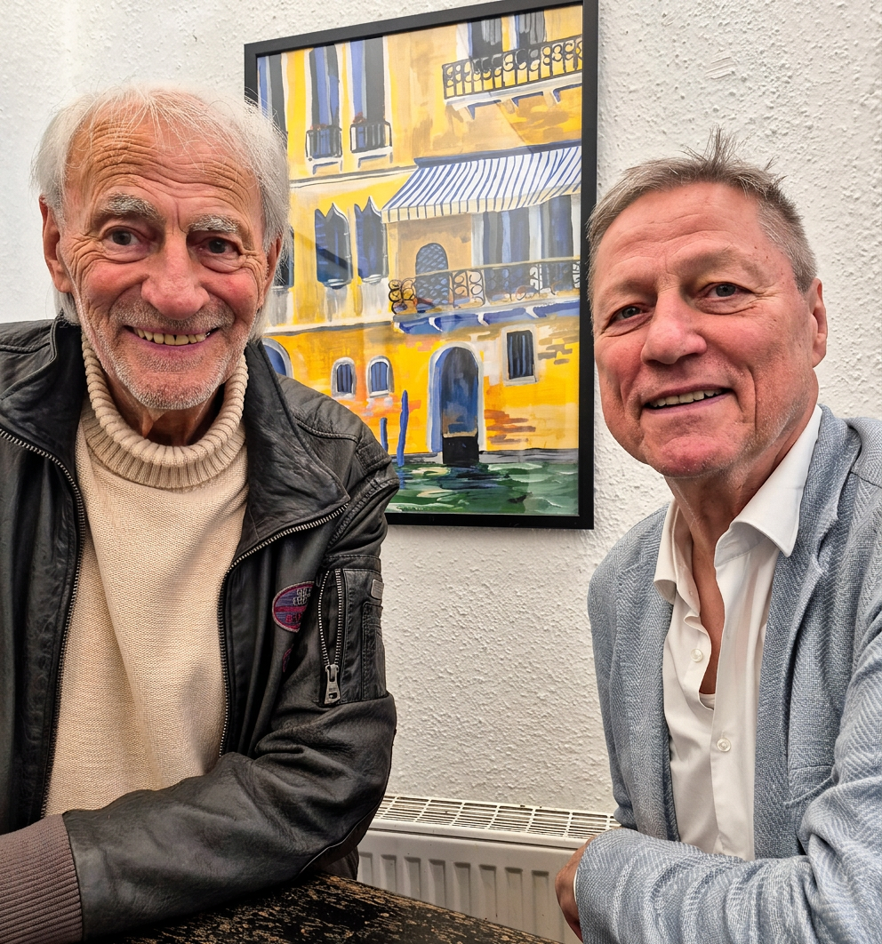 Zwei ältere Männer sitzen an einem Tisch und lächeln in die Kamera, während im Hintergrund ein gemaltes Bild mit einem venezianischen Gebäude zu sehen ist.