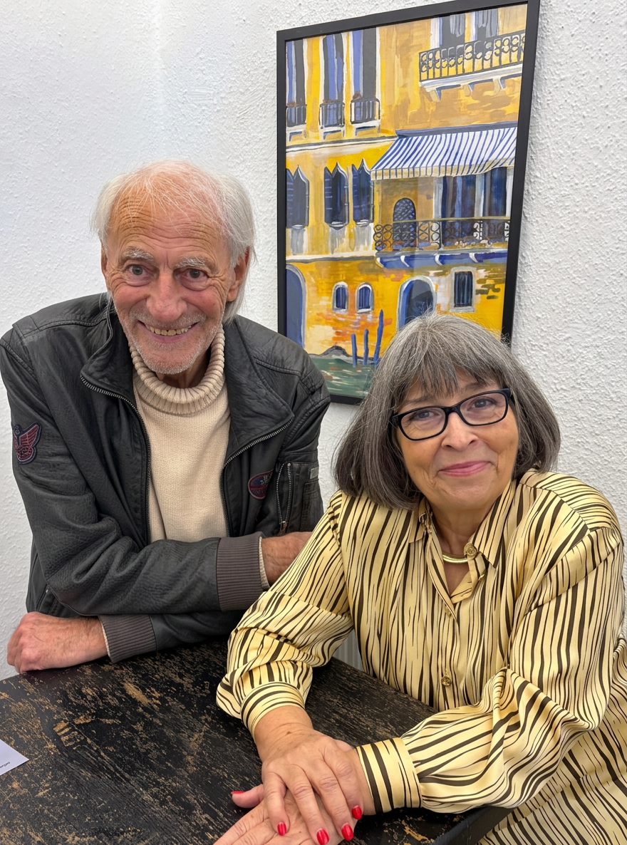 Ein älteres Paar lächelt zusammen an einem Tisch. Der Mann trägt eine schwarze Jacke und einen beigen Pullover, die Frau trägt eine gestreifte Bluse. Im Hintergrund ist ein buntes Kunstwerk an einer Wand zu sehen.