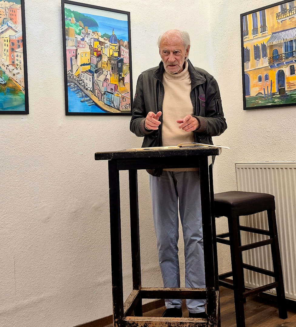 Ein älterer Mann steht an einem Tisch und spricht, während er mit den Händen gestikuliert. Im Hintergrund sind farbenfrohe Gemälde von einer Stadtlandschaft zu sehen.