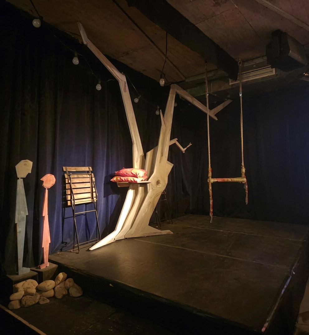 Bühne mit einem außergewöhnlichen Baumkunstwerk, einem Holzstuhl, zwei stilisierten Holzfiguren und einer Schaukel, umgeben von dunklem Vorhang.